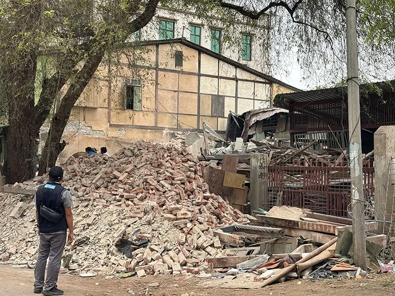 Plan International staff member by fallen building which is a pile of bricks and rubble
