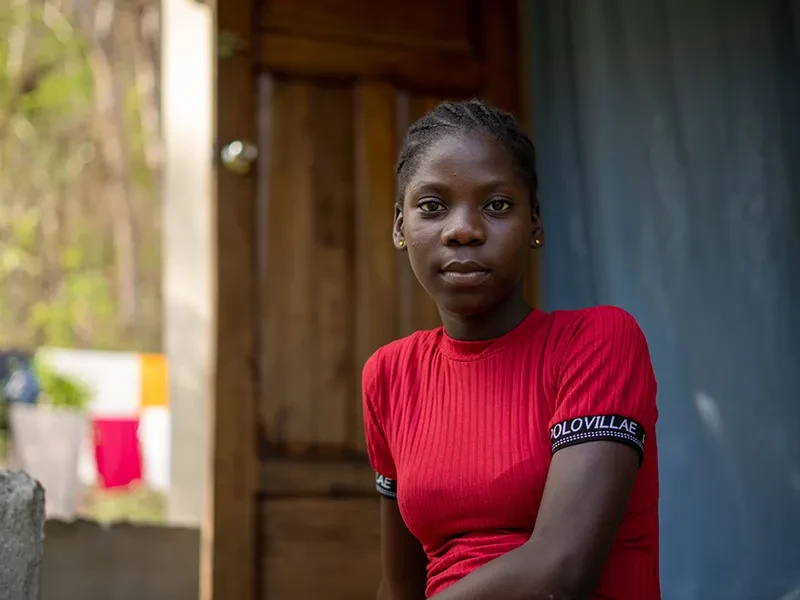 Sofiana, 13, from Haiti, sits outside her family's home. She is wearing a red top and looking at the camera.