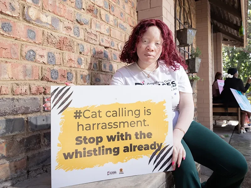Sisasenkosi, 18, from Zimbabwe, sits next to a sign protesting cat calling and harrassment.