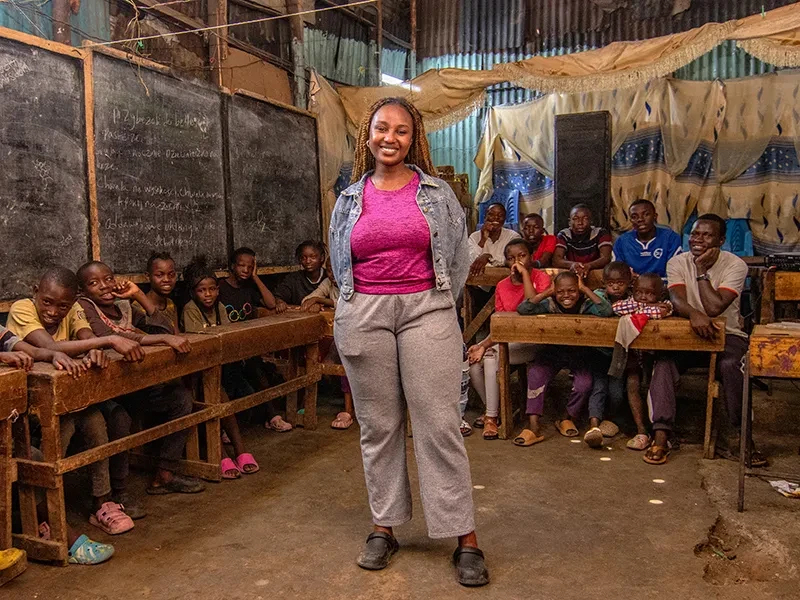 Latifah stands in the middle of a classroom. She is surrounded by young girls and boys who are seated at desks.
