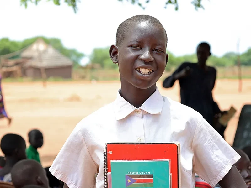 Monica, 14, wears a white shirt. She is smiling and holding her school textbooks.