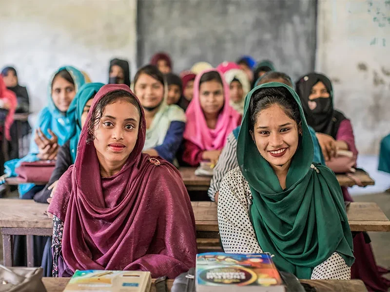 Shunhana and Sheemla sitting at classroom desk, with other students sitting around them out of focus.