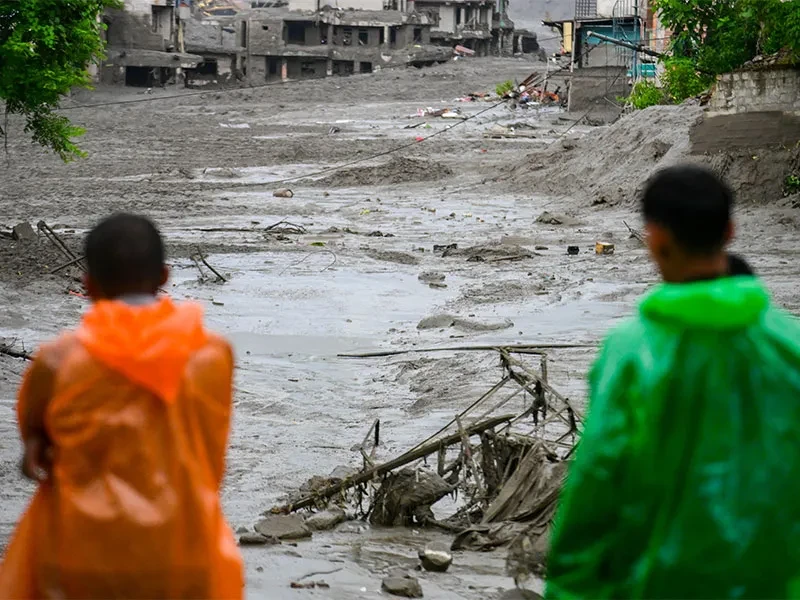 Backs of two people in rain ponchos looking at flood and mudslide with destroyed buildings and debris in view