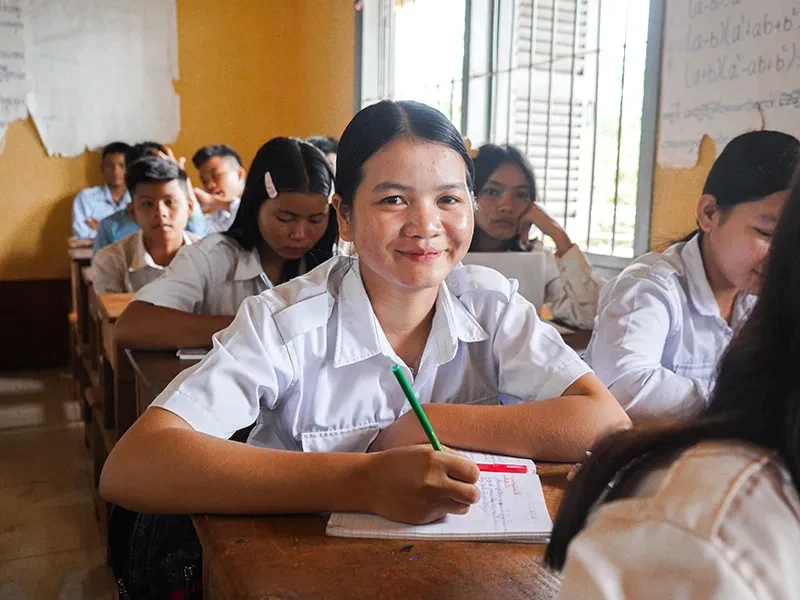 Phgnon, 16, is sitting in a classroom, writing in a notebook. She is surrounded by other students.