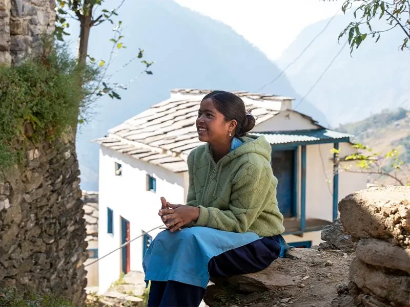 Kokila sitting outside with house and mountains in background.