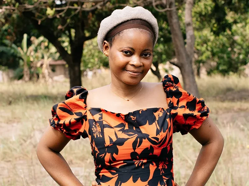 Fatmata, 19, is wearing an orange and black dress. She stands in a field with trees behind her.